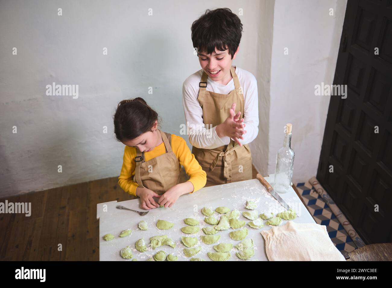 View from above of two diverse kids, a boy and a girl preparing family dinner, standing at floured kitchen table and modeling dumplings or Ukrainian v Stock Photo