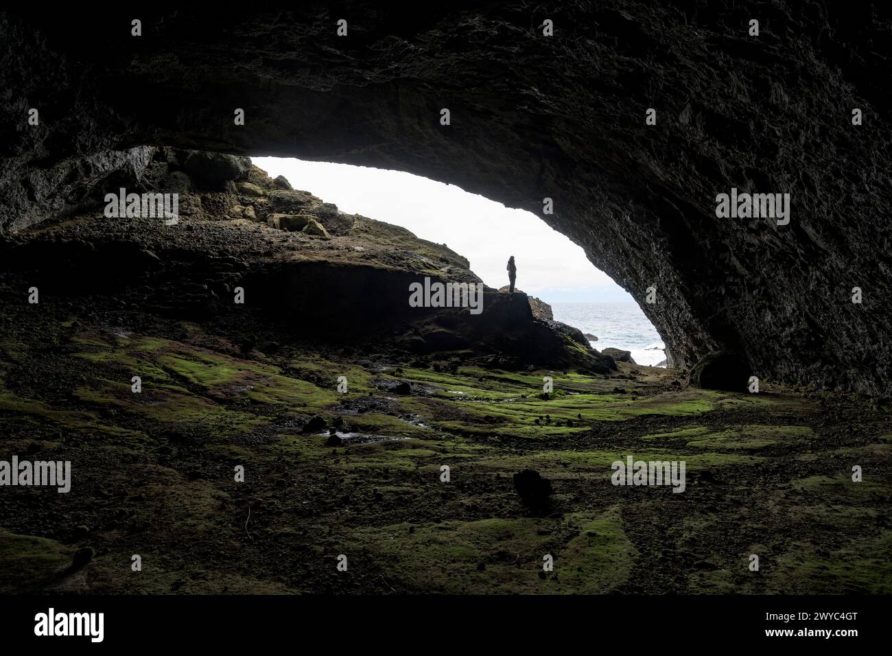 A person stands at the mouth of a large sea cave, silhouetted against the bright sky and rugged coastline Stock Photo