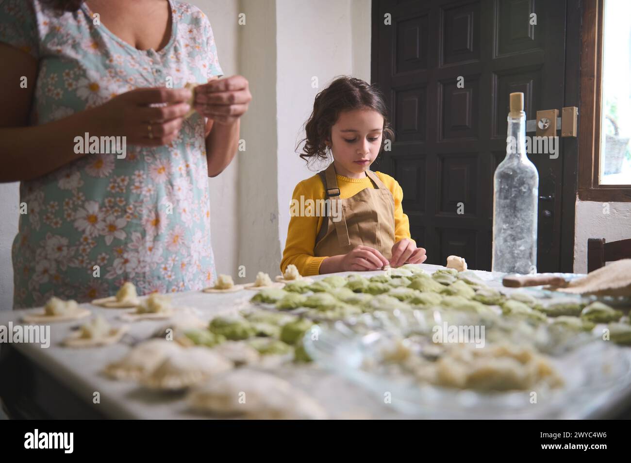 Lovely child girl in beige chef apron, molding dumplings with mashed potato filling, standing at floured table near her mother, helping her in cuisine Stock Photo