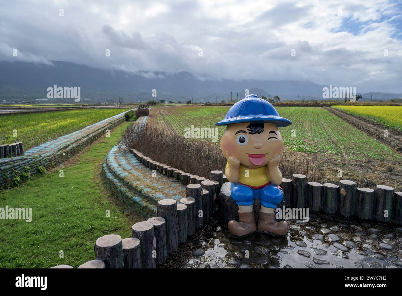 A childlike figure sits on logs gazing over a verdant farmland under a cloudy sky in Mr. Brown Avenue farmland Stock Photo