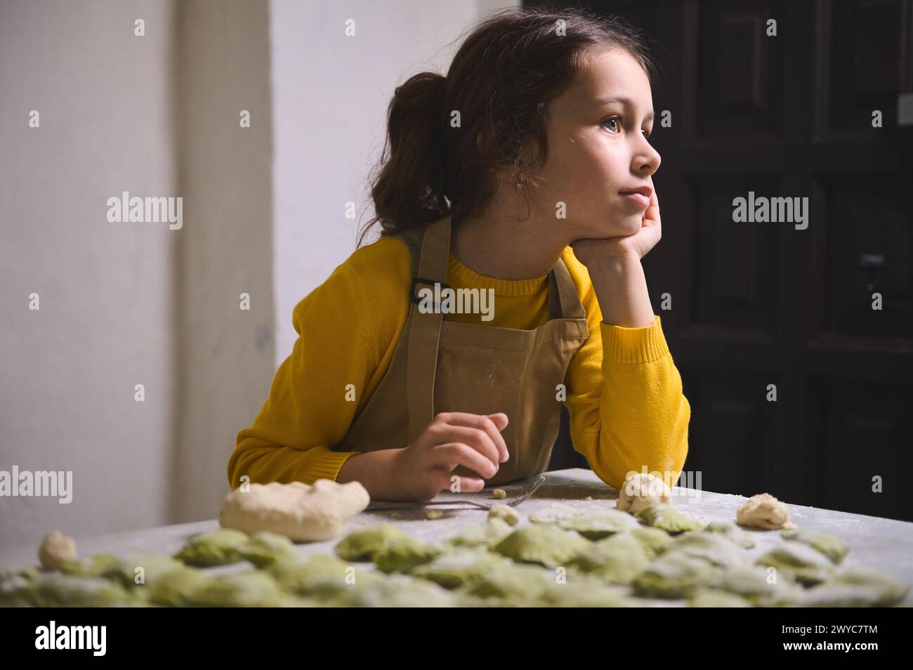 Authentic portrait of Caucasian beautiful little girl 6 y.o, in beige chef apron, dreamily looking away, standing at floured table with molded dumplin Stock Photo