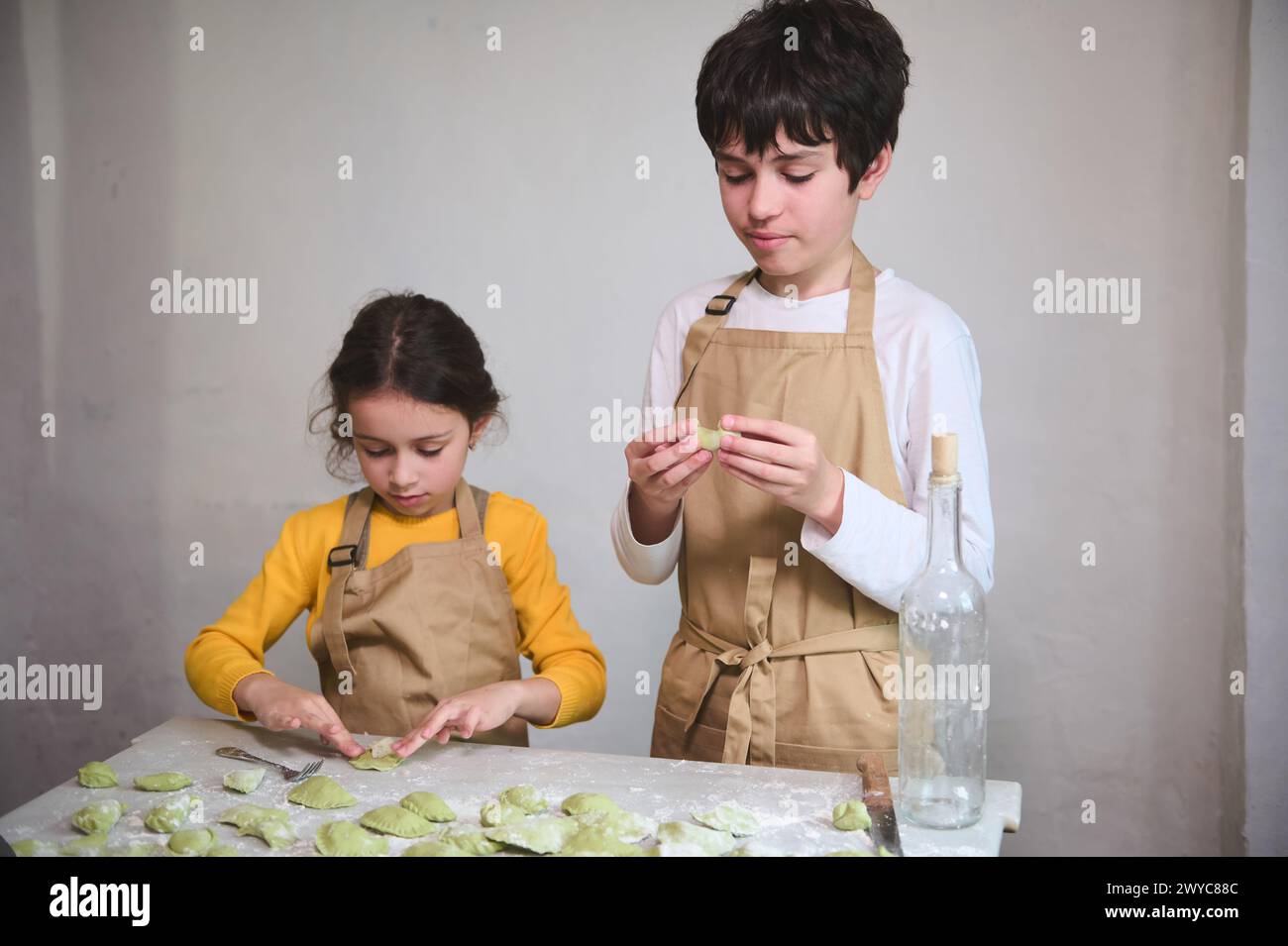 Cooking class for kids. Two diverse kids, a boy and a girl preparing family dinner, standing at floured kitchen table and modeling dumplings or Ukrain Stock Photo