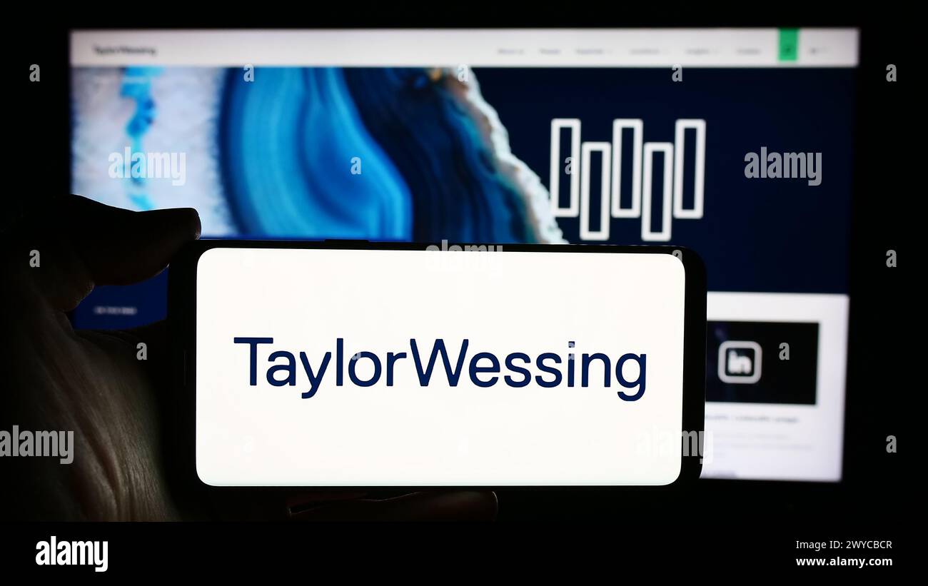 Person holding cellphone with logo of international law company Taylor Wessing LLP in front of business webpage. Focus on phone display. Stock Photo