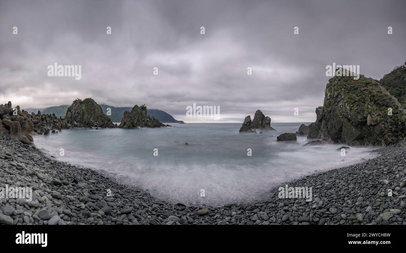 A panoramic view of a serene seascape with rocky formations under a dynamic, overcast sky Stock Photo