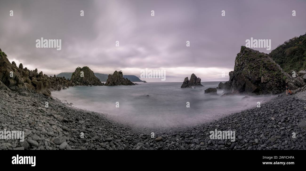 A panoramic view of a serene seascape with rocky formations under a dynamic, overcast sky Stock Photo