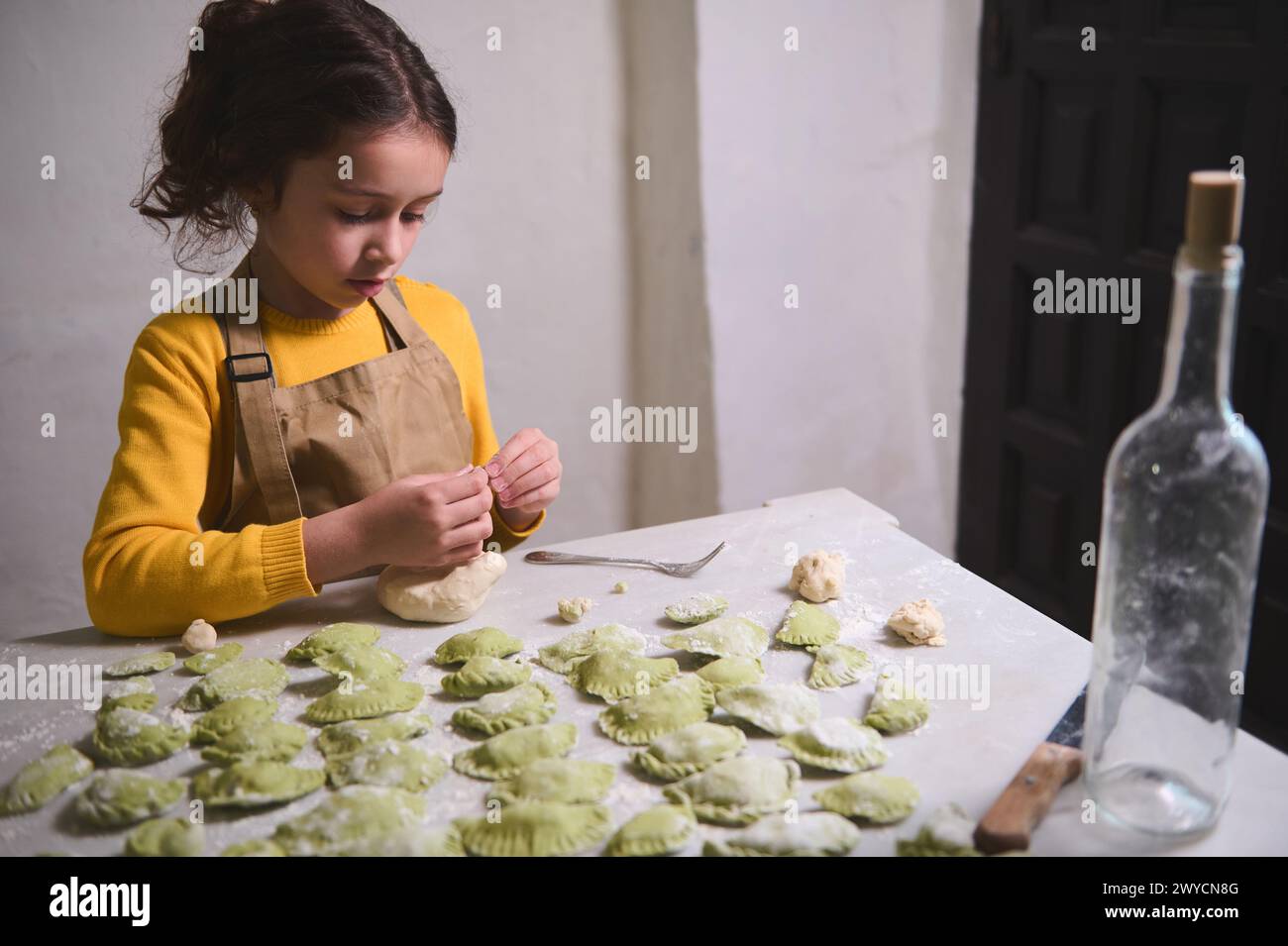 Caucasian little girl in beige chef apron, molding dumplings, standing at floured kitchen table. Kid girl helping her mom cooking dinner at home Stock Photo
