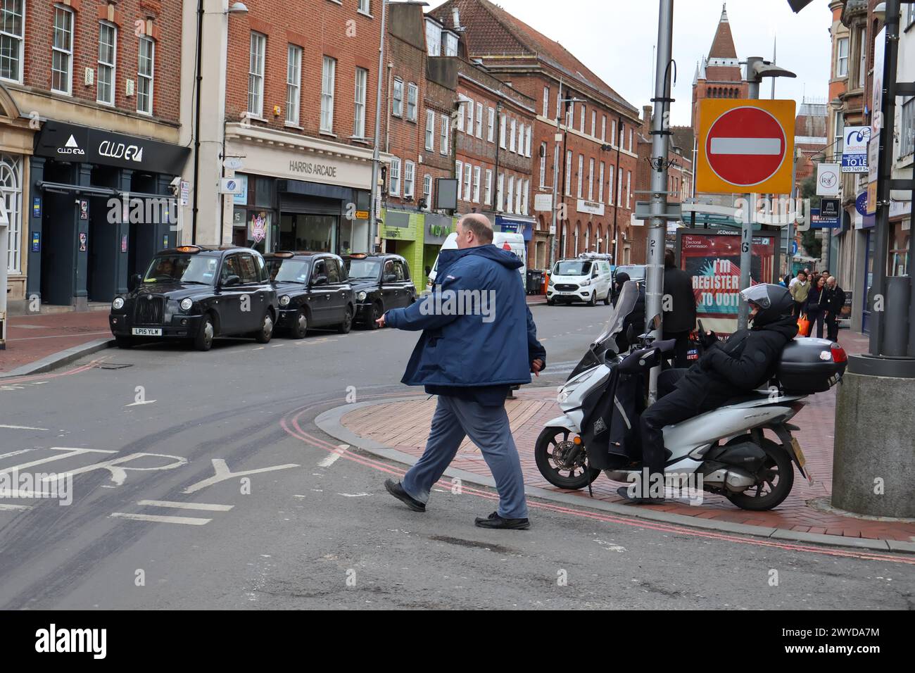 Pedestrian walking across the road at the taxi rank as a delivery rider lays back relaxing on his motorcycle scooter. Stock Photo