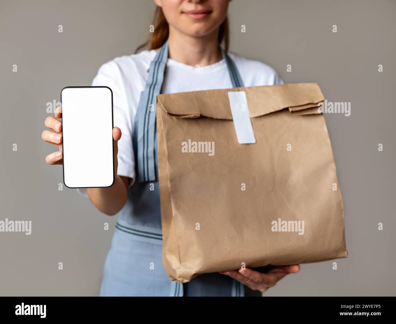 Mock-up banner for mobile app of order and delivery food. Waitress holds mobile phone with empty screen and paper bag for takeout meal in her hands. Stock Photo