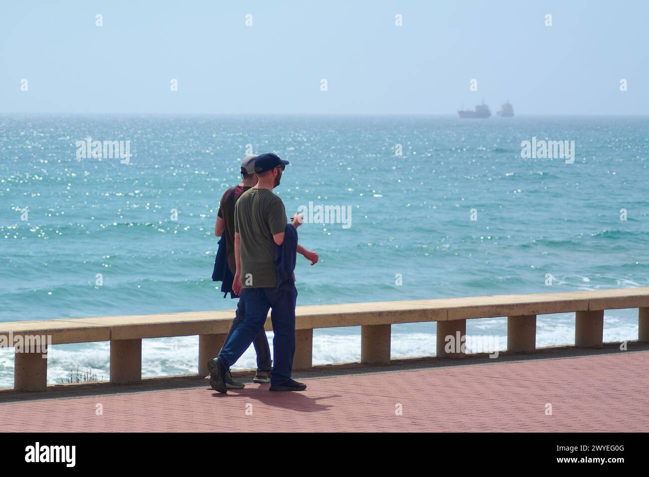 Tarragona, Spain - April 6, 2024: Two people enjoying a sunny boardwalk, with the blue sea and a boat in the distance, reflecting the tranquility of t Stock Photo