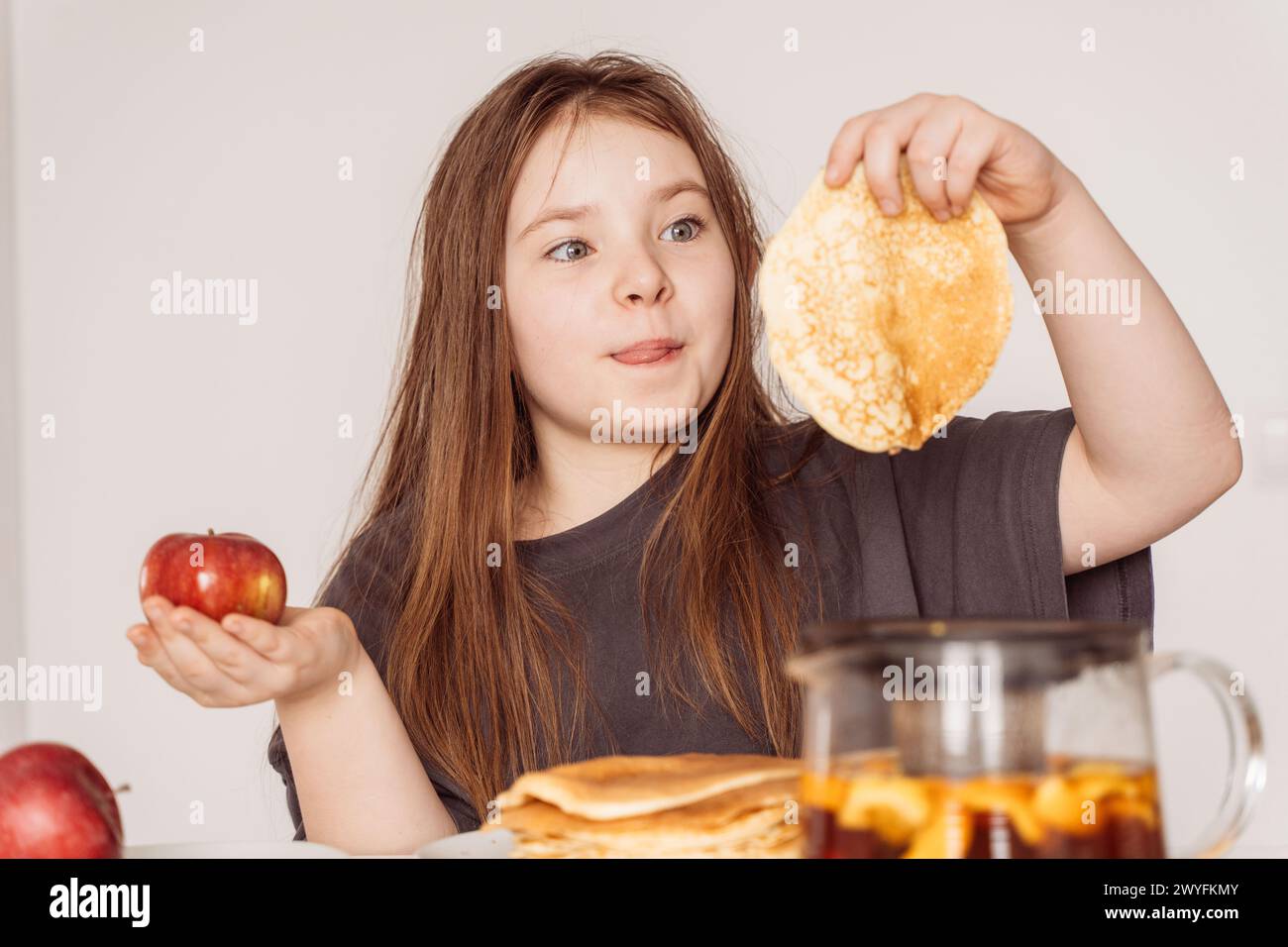 Little teenage girl holding an apple in one hand in the other pancake. The girl chooses between an apple and a pancake for breakfast, Looks licking her lips at the pancake. Food selection concept. High quality photo Stock Photo