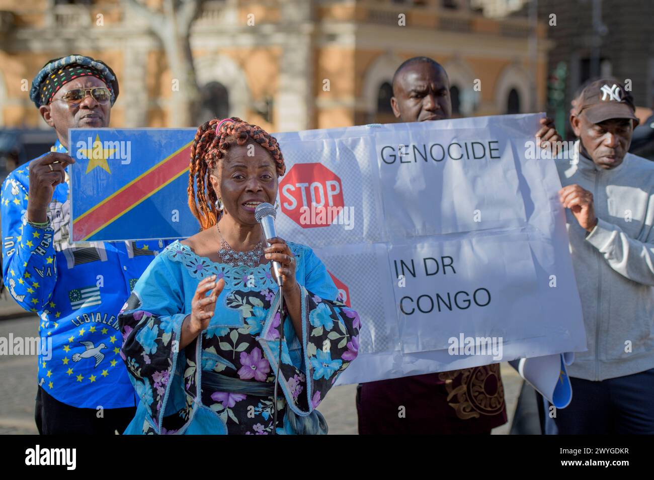 April 6, 2024, Rome, Italy: Protesters display the Congo flag and a sign reading 'stop the genocide in the DR of Congo' while a woman in traditional African dress speaks into the microphone during the demonstration to express solidarity with the Congolese people, to ask for a 'stop to the ongoing genocide' and an end to the exploitation of the African continent organized by some associations of the African communities of Europe together with the Communist Network and the Hands Off from Africa movement in Rome.According to the organizers, the 30 years of armed conflict financed by the Western w Stock Photo