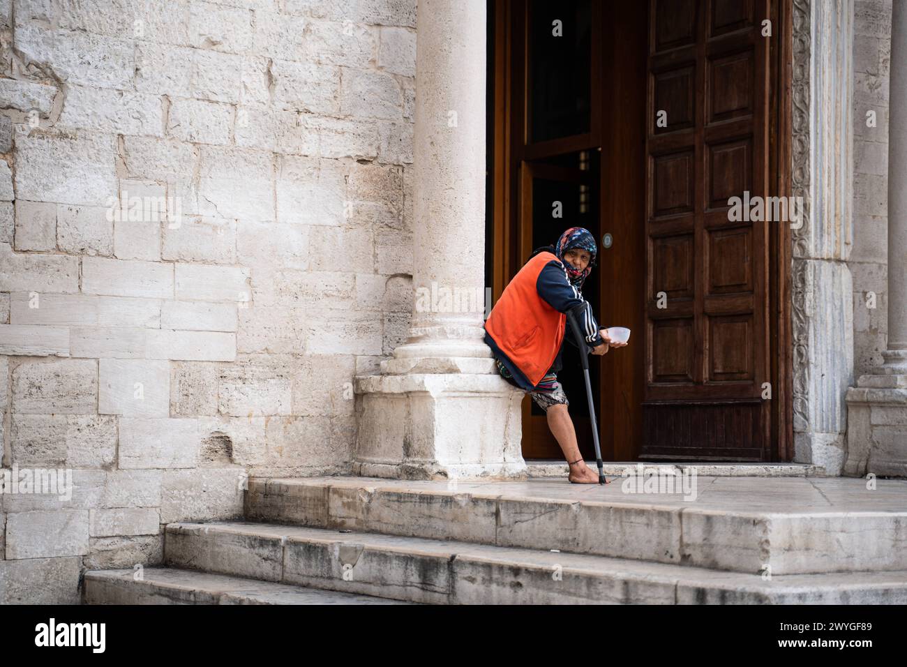Bari, Italy - September 22, 2023: An old woman on crutches begs for money in front of a church in Bari, Italy *** Eine alte Frau auf Krücken bettelt vor einer Kirche in Bari, Italien um Geld Stock Photo