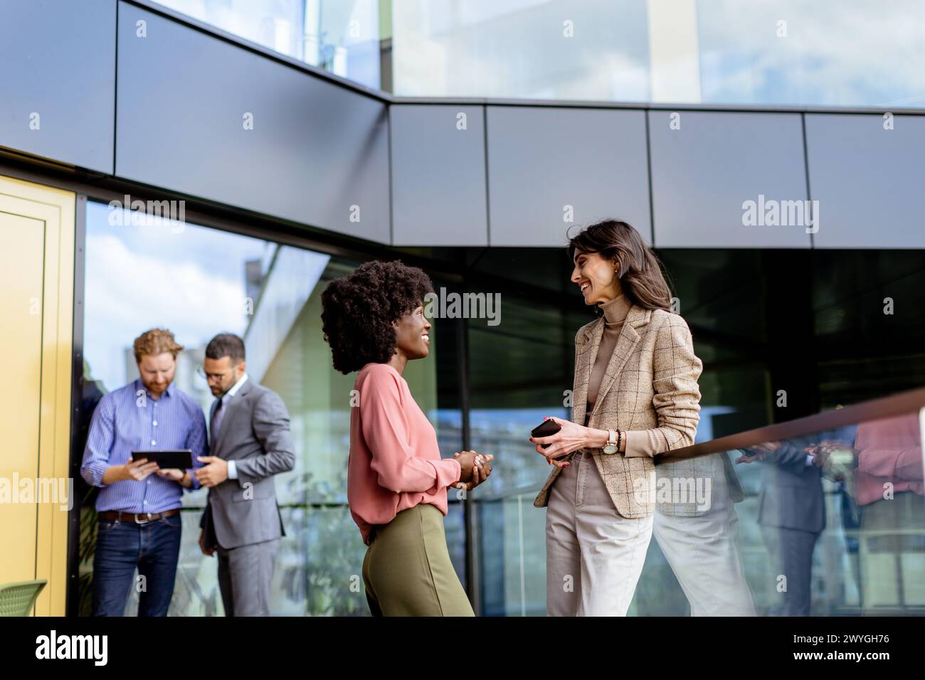 Two female multiethnic colleagues engage in a friendly chat outside an office, with others working in the background Stock Photo