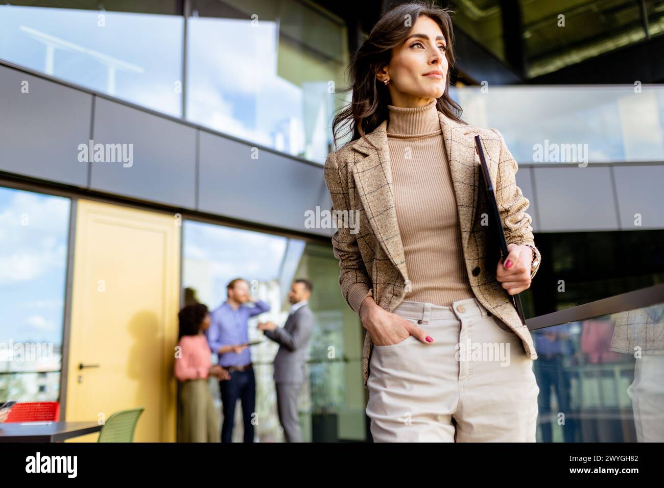 Stylish professional strides forward, glancing to the side, as colleagues converse in the background Stock Photo