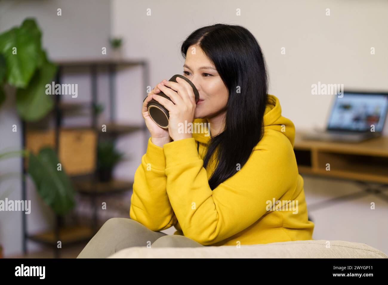 A woman in a yellow hoodie is drinking coffee. She is smiling and she is enjoying her drink Stock Photo