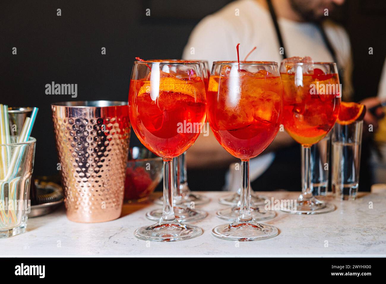 Refreshing Aperitif Spritz cocktails with ice and orange slices, served in wine glasses on a marble bar counter, with a bartender in the background. Stock Photo