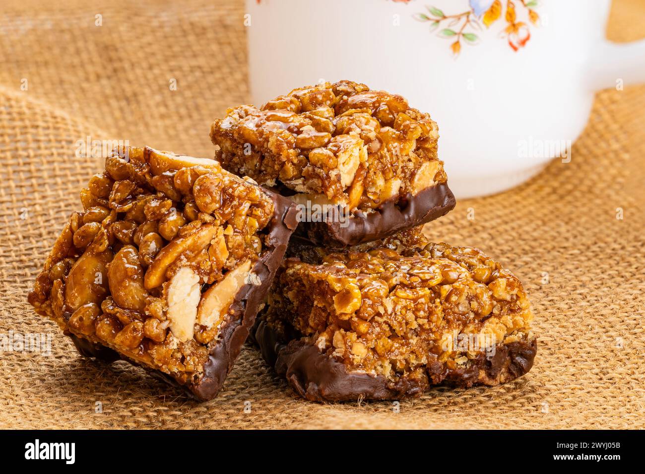 Closeup view of crispy homemade peanuts, almond and chocolate mini protein bars and white coffee cup on sack cloth. Stock Photo