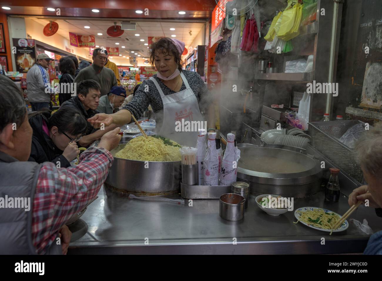 A woman serves noodles at a busy market stall, taking money from a customer in a bustling environment Stock Photo