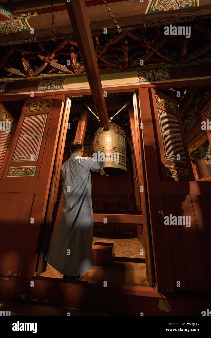 A monk in traditional attire ringing a large bell in Baoan temple during Chinese New Year celebrations Stock Photo