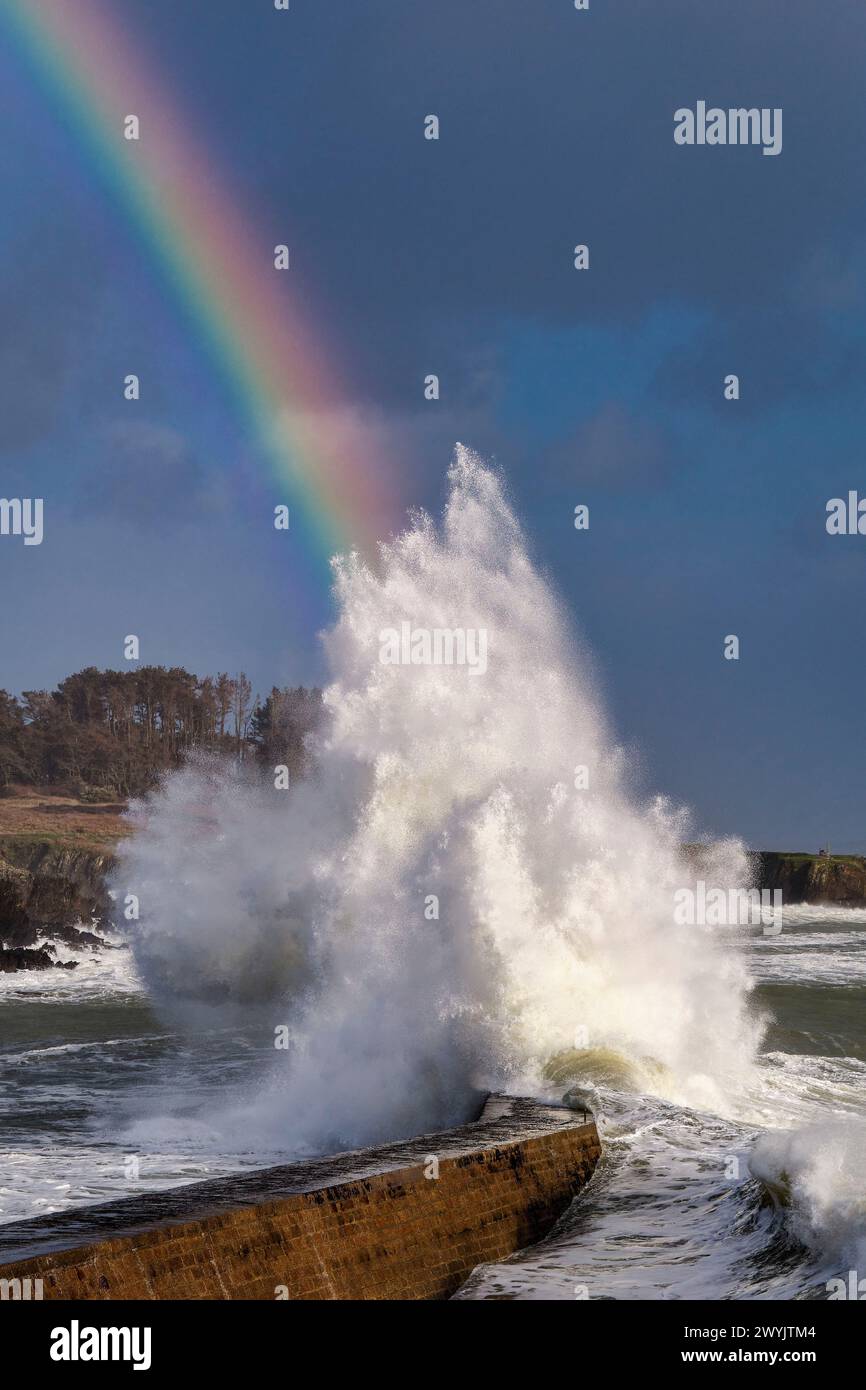 France, Finistère, Clohars-Carnoet, Doelan, rainbow over a wave rising on the harbour jetty during a winter storm Stock Photo