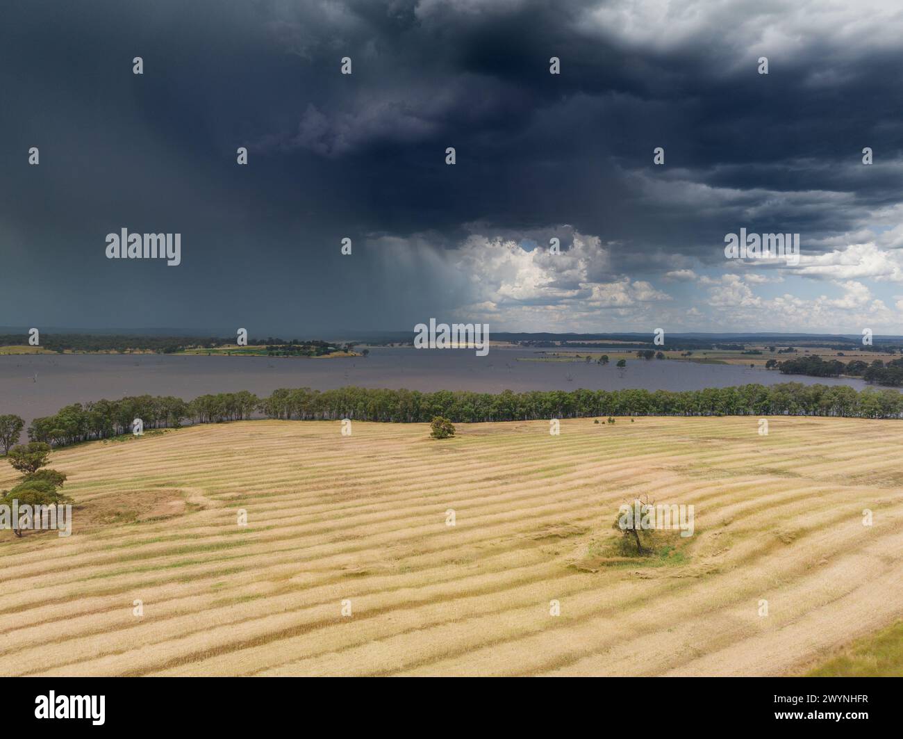 Aerial view of rain falling from dark storm clouds moving over a reservoir behind a dry harvested field at Joyces Creek in Central Victoria, Australia Stock Photo