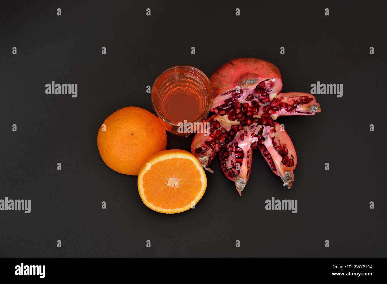 A glass of a mixture of tropical fruits on a black background, next to a half of a ripe orange and a broken pomegranate. Top view, flat lay. Stock Photo