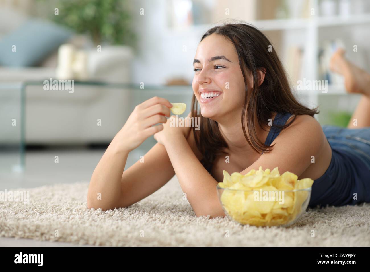 Happy woman eating potato chips lying on a carpet at home looking at side Stock Photo