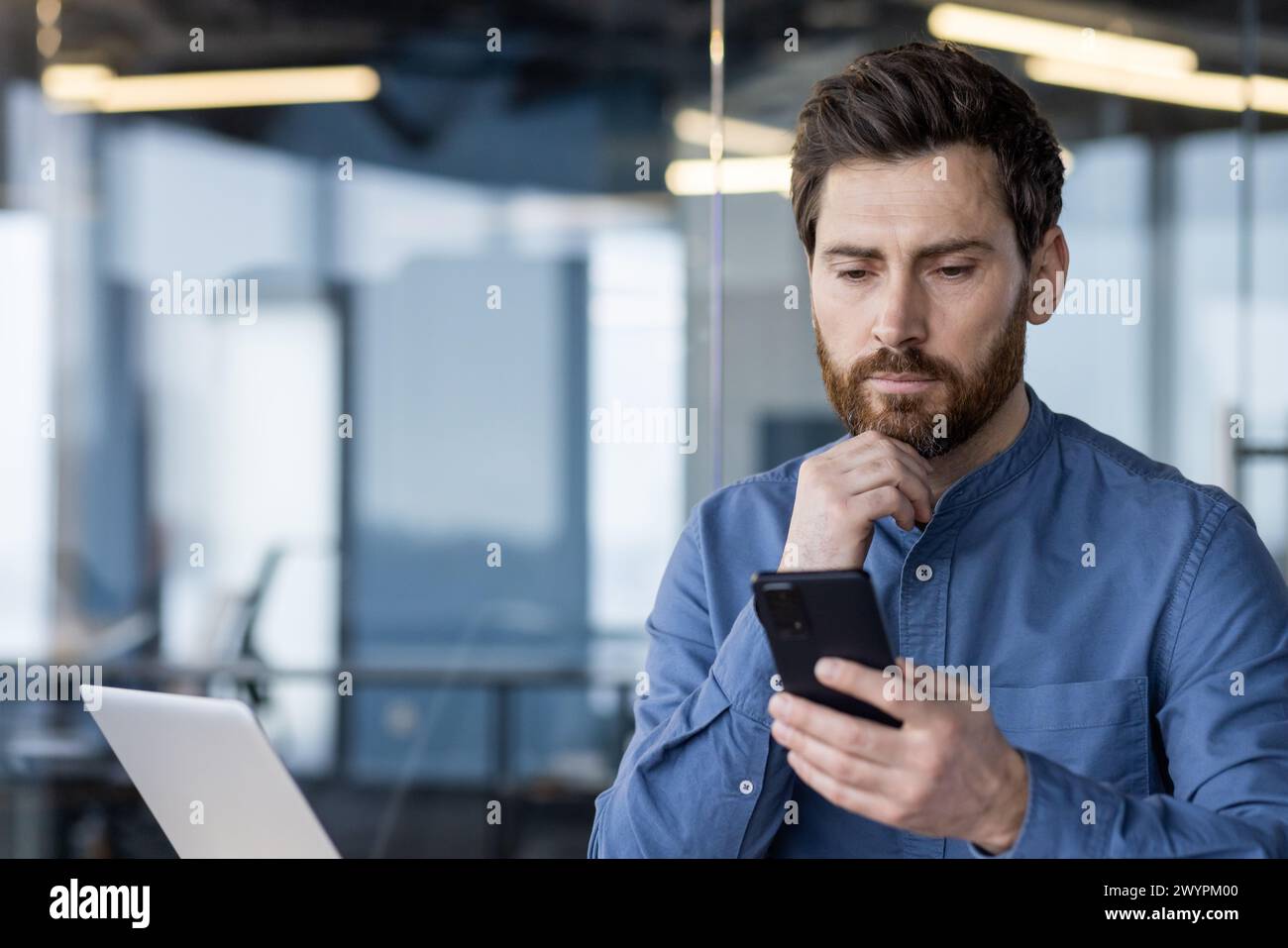 Professional businessman in a modern office setting, intently using a smartphone while working on a laptop. Stock Photo