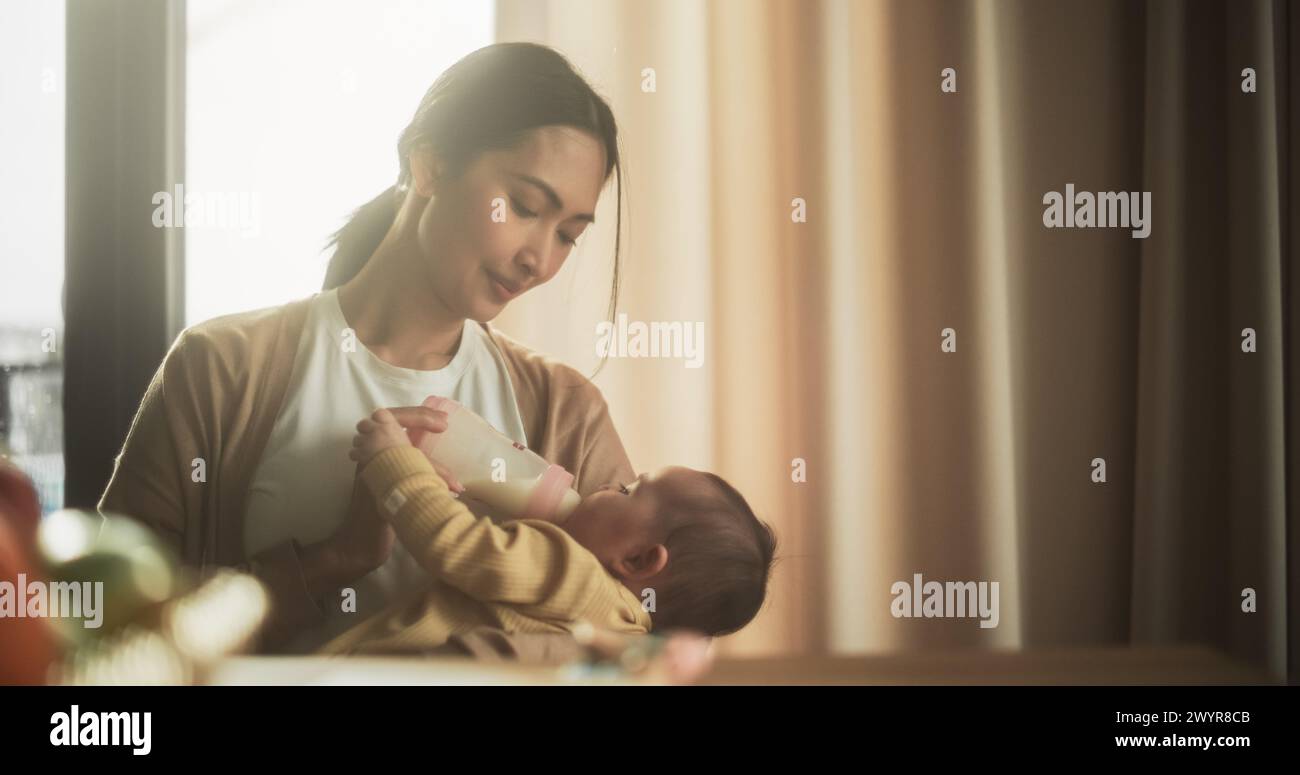 Portrait of Beautiful Asian Mother Feeding Milk to her Infant Using a Baby Bottle at Bright Home. Young Woman New to Motherhood Bonding with her Child and Enjoying an Affectionate Family Moment. Stock Photo