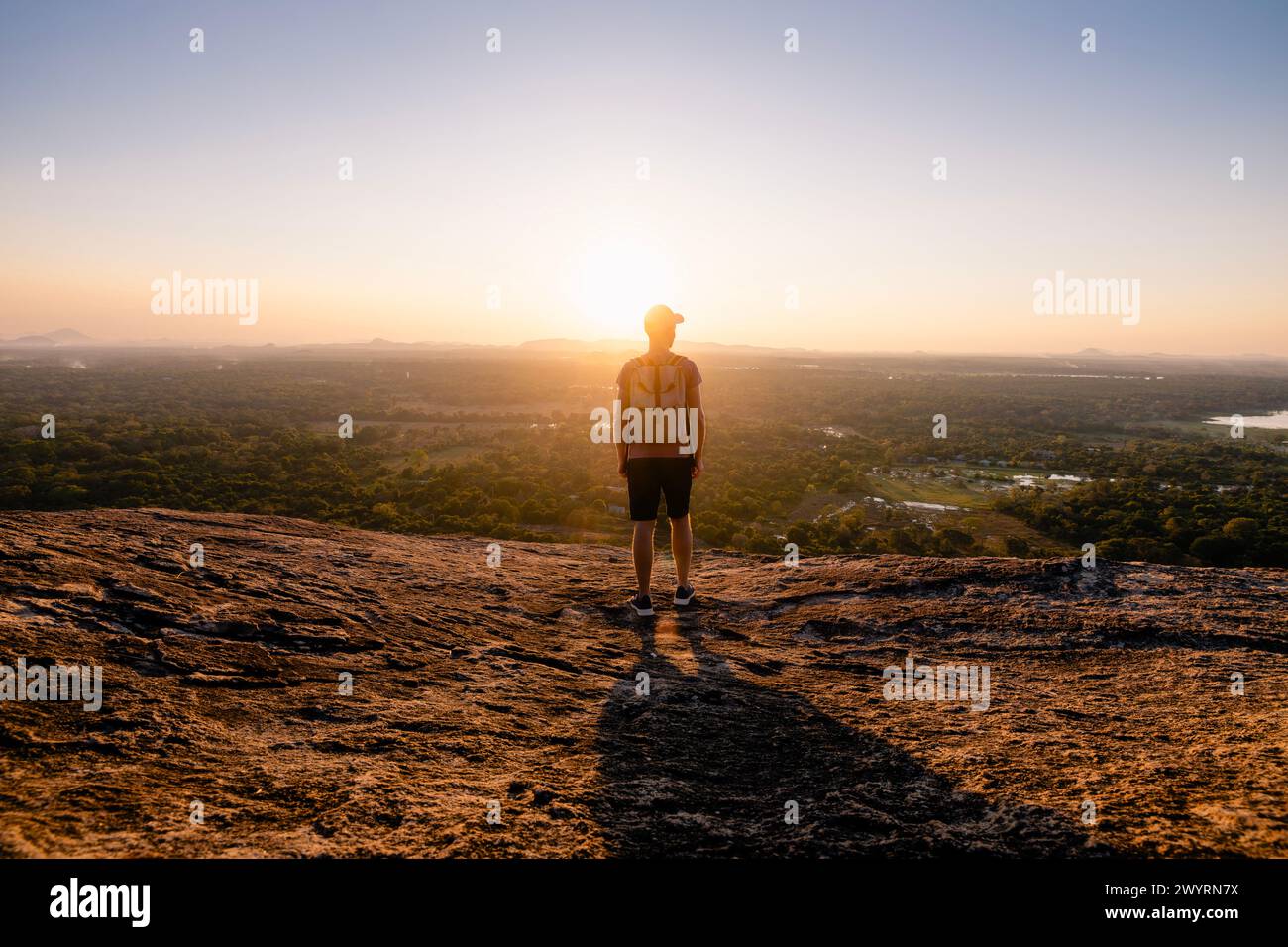 Rear view of man with backpack on rock overlooking sunset over tropical landscape. Travel in Sri Lanka. Stock Photo