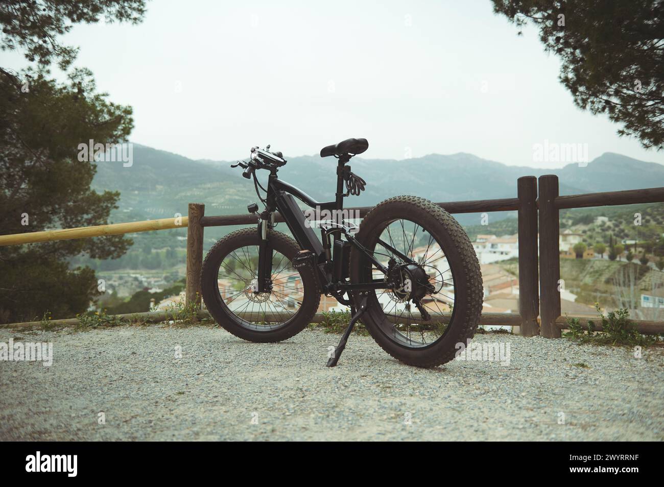 A full size shot of a modern black electric motor bike on the countryside road, parked by a wooden fence, against mountains background in the nature. Stock Photo