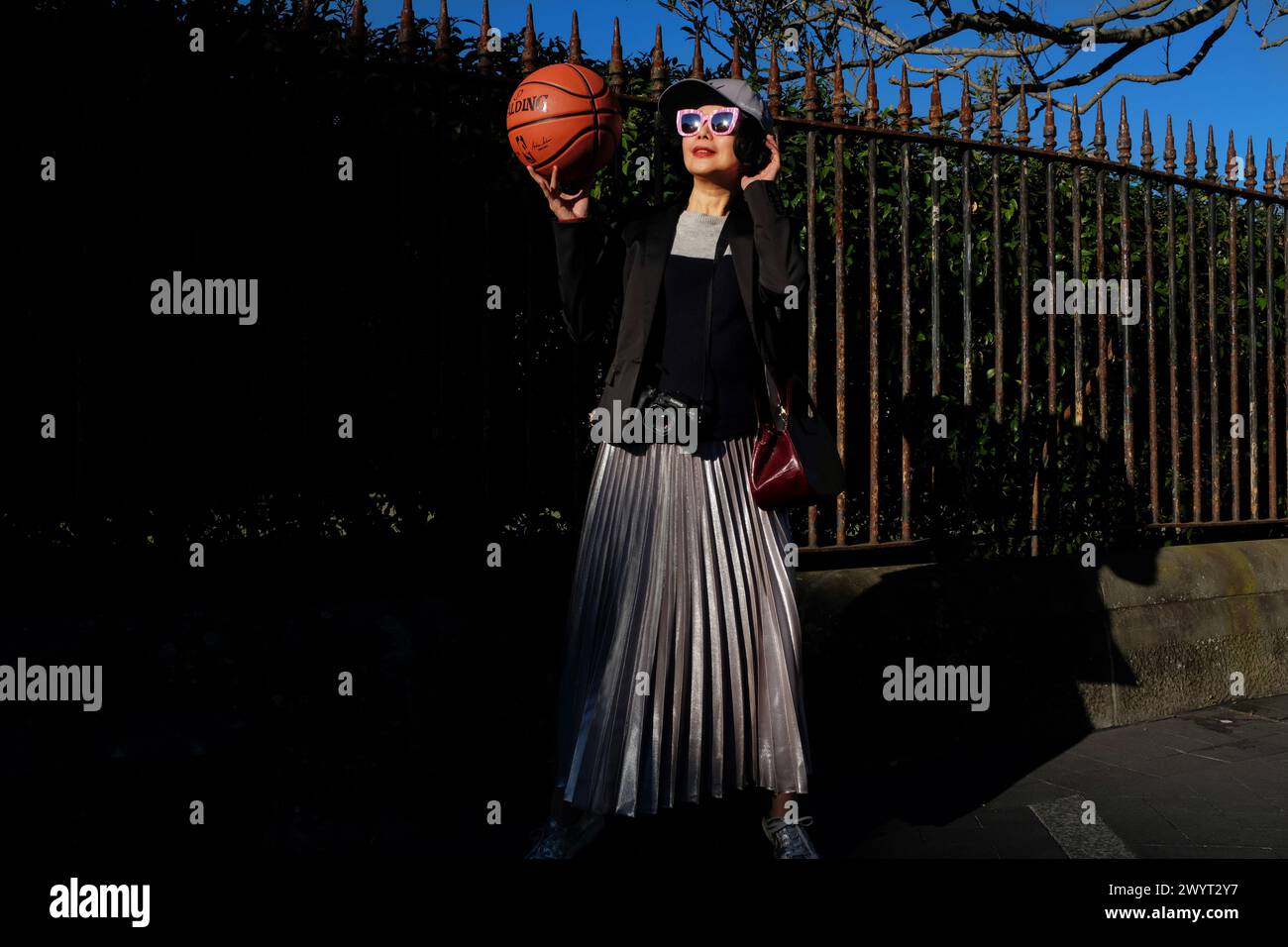 Face of a woman in pink sunglass wearing a baseball cap, holding a Spalding basketball in afternoon light outside a tall iron palisade fence Stock Photo