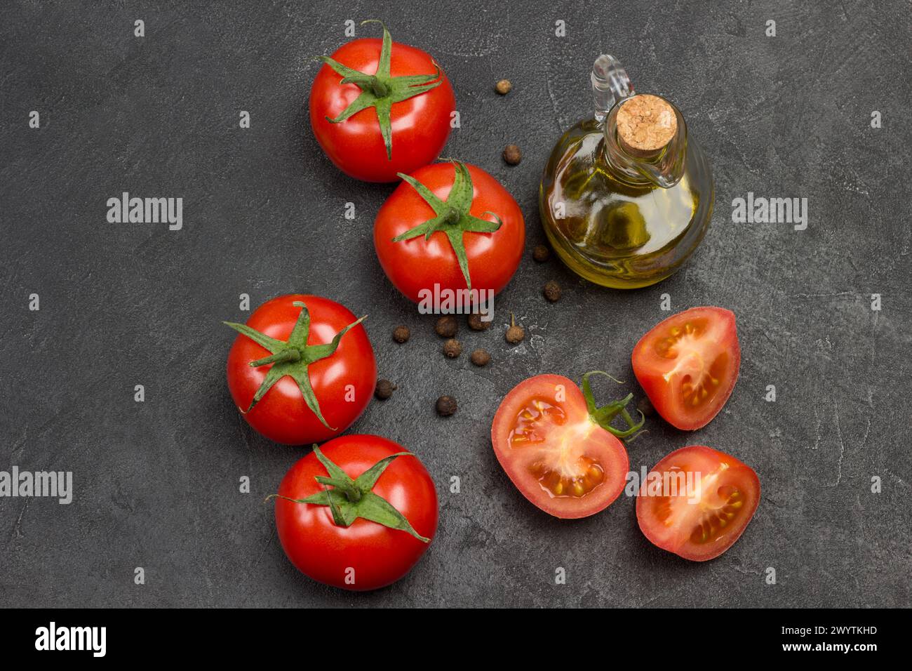 Whole tomato and chopped tomato wedges, allspice on table.  Bottle of olive oil and sprig of rosemary.  Black background. Top view. Stock Photo