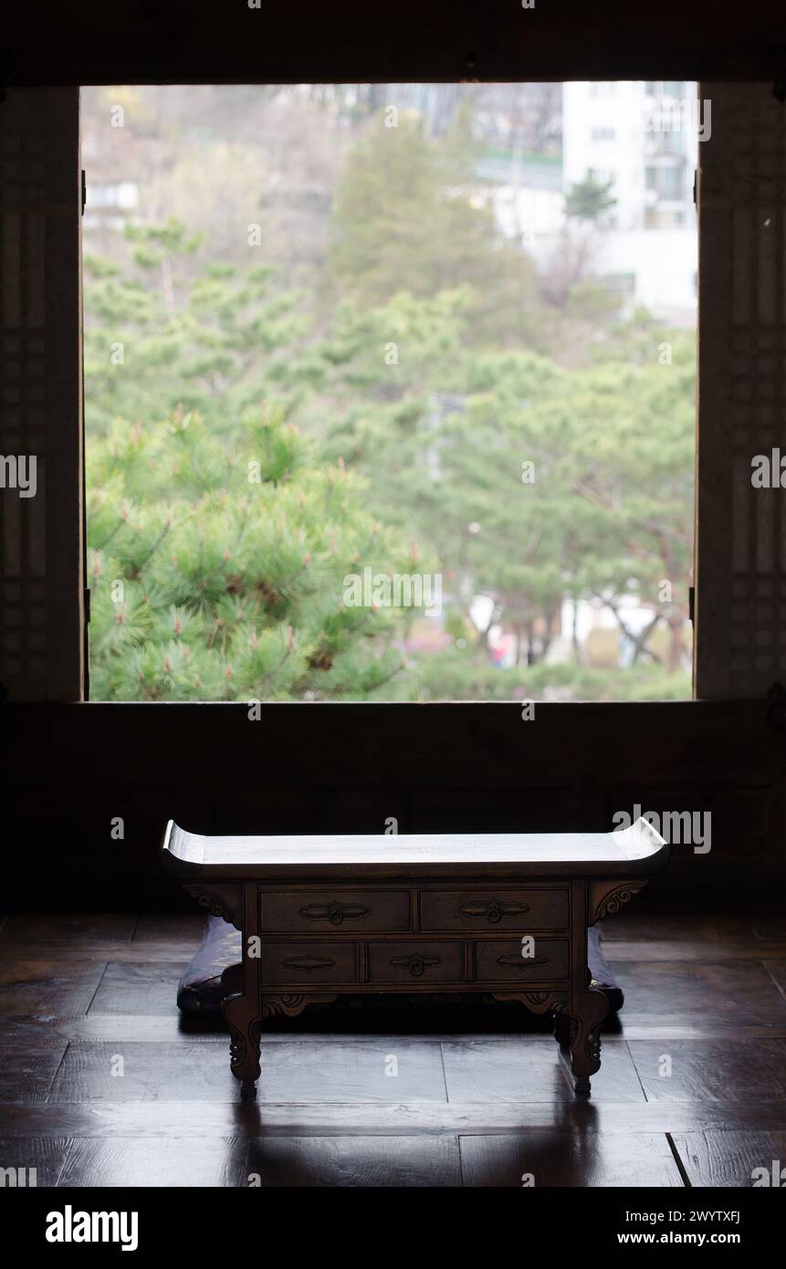 A wooden table with a bench sits in front of a window. The bench is empty and the table is covered with a blanket. The room is dimly lit and the view Stock Photo