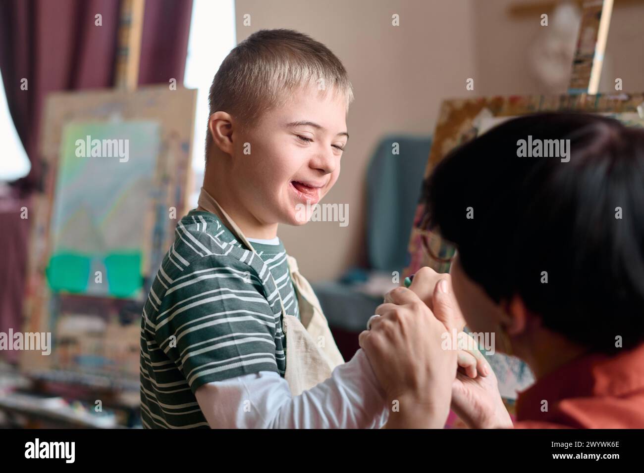 Portrait of young boy with Down syndrome laughing cheerfully enjoying art class with parent or teacher assistant Stock Photo