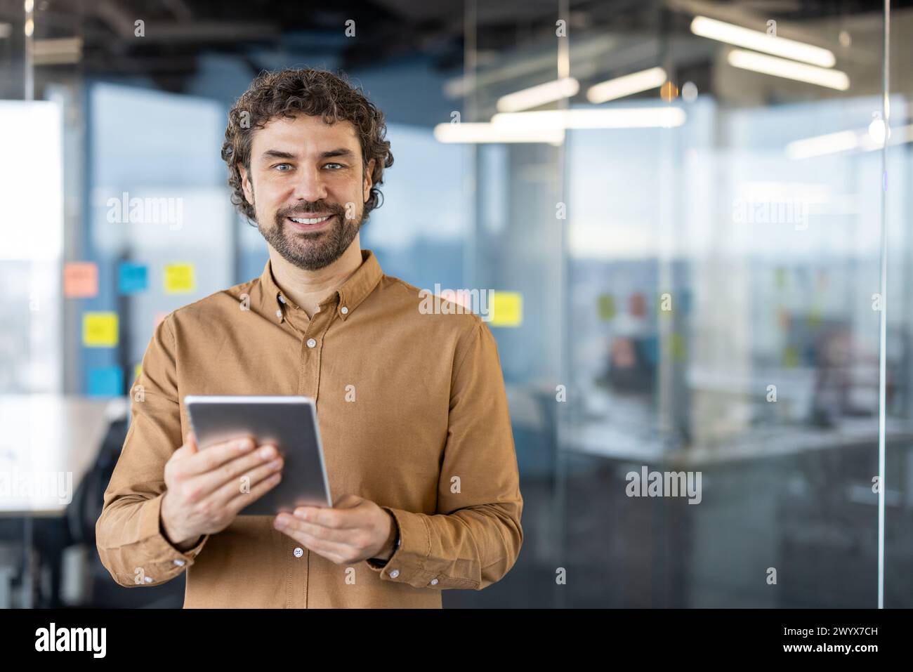 Portrait of a mature Hispanic businessman smiling confidently while holding a digital tablet in an office setting with glass walls. Stock Photo