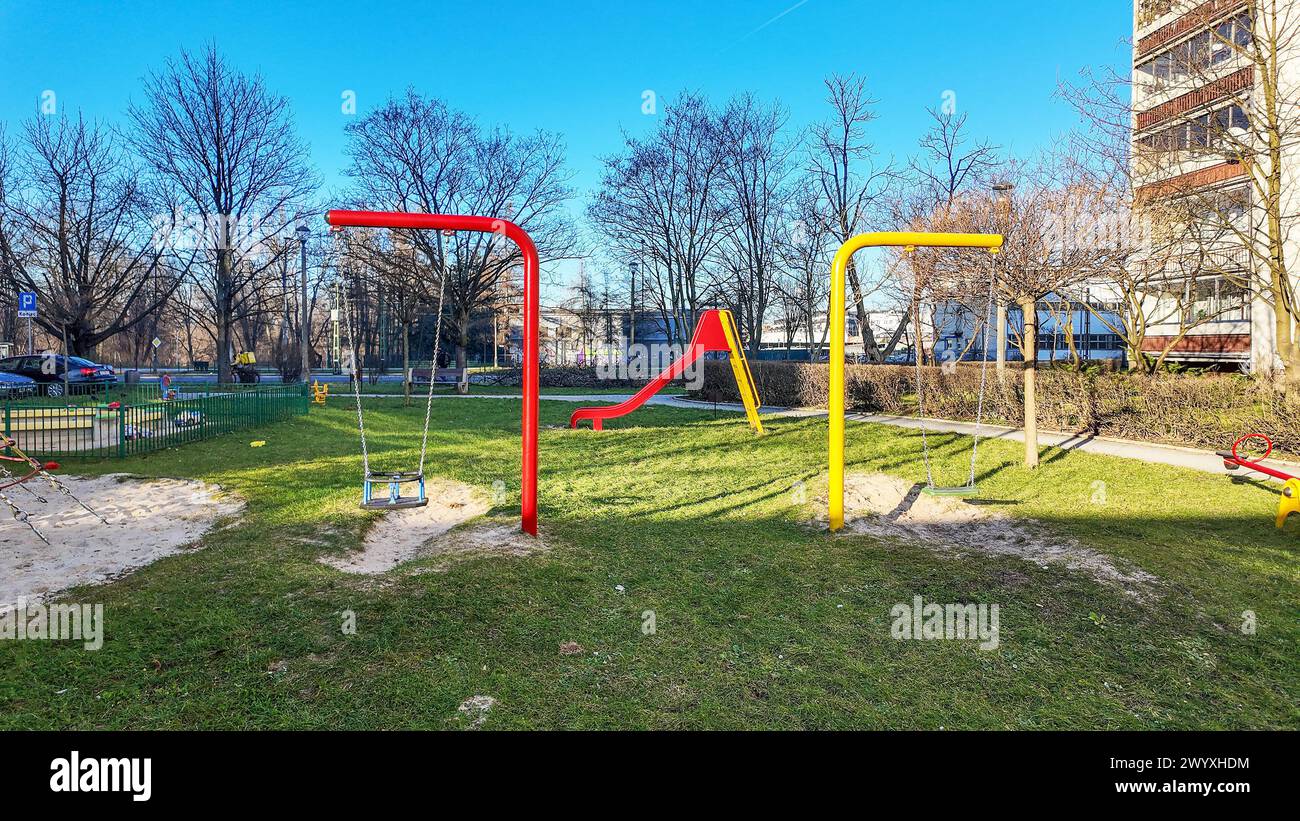 Playground for children with two swings and a slide in yellow and red colors. Sandbox on the left. Urban recreation and play area between blocks of fl Stock Photo
