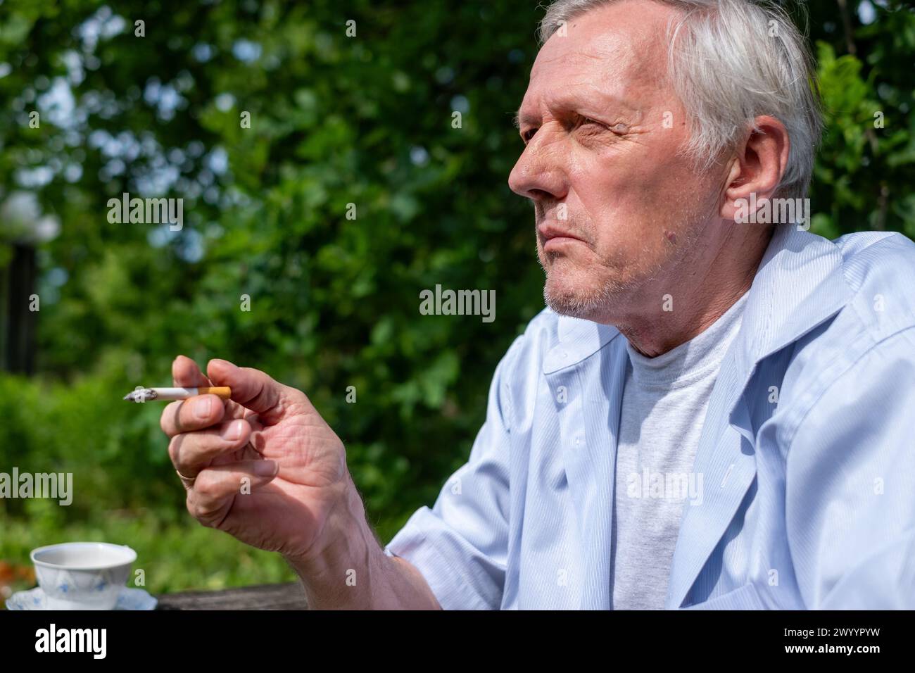 Deep in thought, a senior individual, elderly man, pauses for a smoke break outside, amidst the freshness of a vibrant garden, addiction at an old age Stock Photo