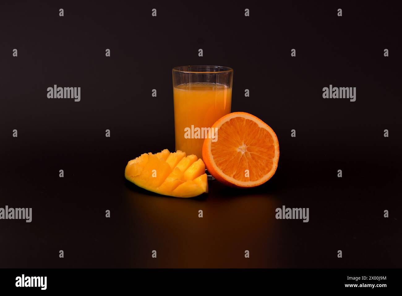 A glass glass of a mixture of tropical fruit juice on a black background, next to pieces of mango and ripe orange. Close-up. Stock Photo