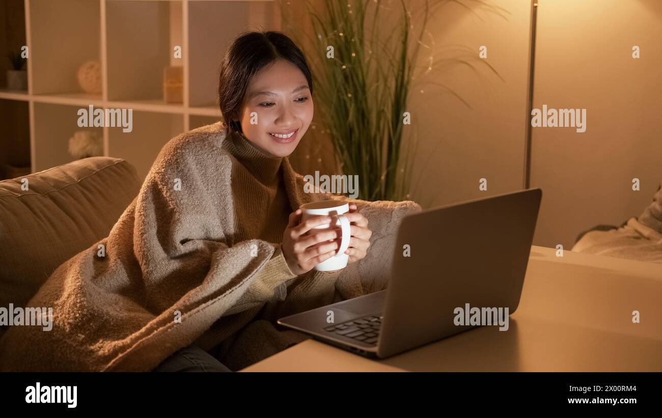 Hygge evening. Home rest. Weekend leisure. Relaxed happy woman watching movie on laptop with hot tea alone on cozy couch. Stock Photo