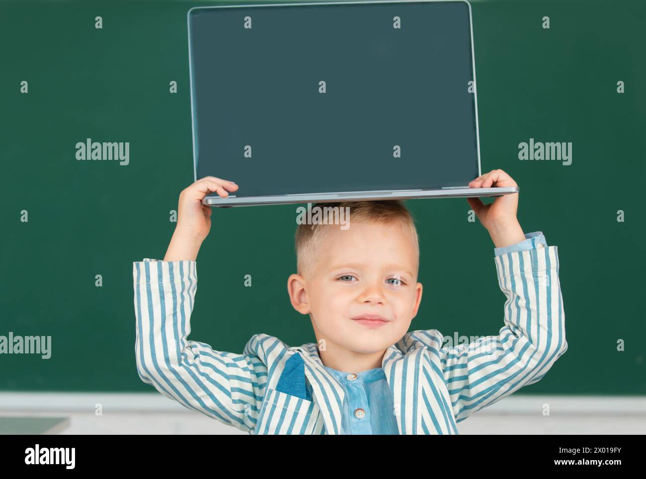 School child holding laptop on head using digital pc laptop in class. Portrait of funny pupil of primary school study programming. Stock Photo