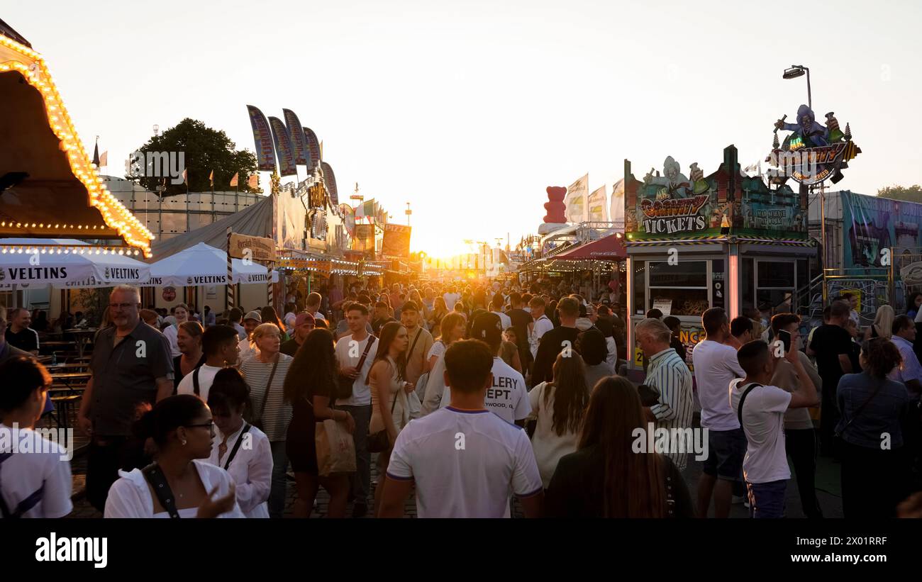Herne, Germany - August.07.2023: View into a street from a funfair with sunset at the end. The crowd strolls past food stalls and the ride 'Mr Gravity Stock Photo