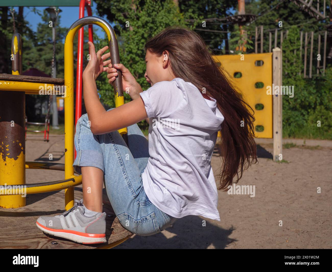 Closeup of a happy smiling teenage girl with long hair in a white t-shirt and blue jeans spinning at a merry-go-round at children playground Stock Photo