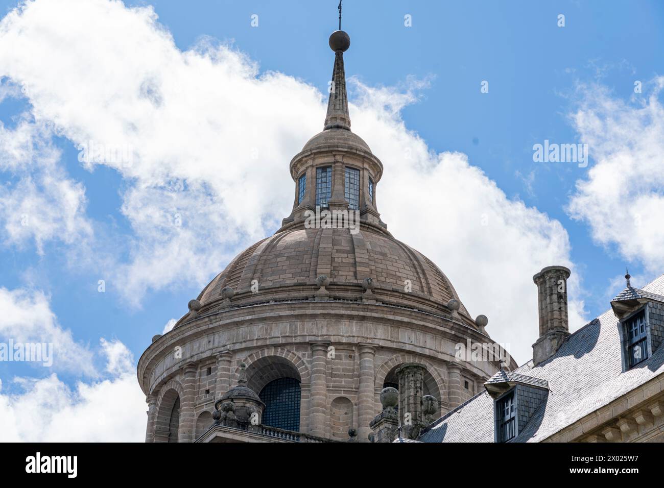 Sculptural details and the iconic dome of El Escorial Monastery stand out against a blue sky with fluffy clouds Stock Photo