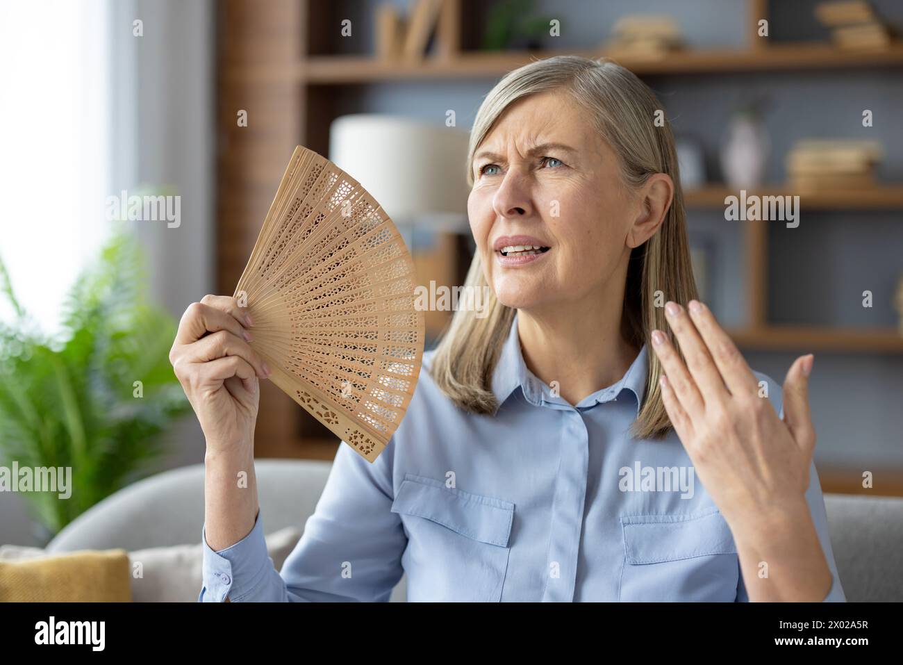 Disturbed woman waving with wooden hand fan to squinted face while sitting in overheated apartment. Exhausted woman suffering from no air conditioner at home during summer temperature increasing. Stock Photo