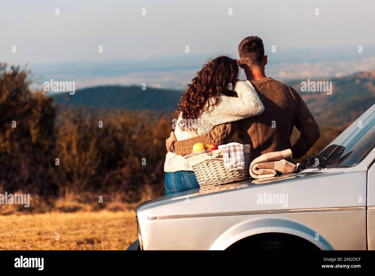 Rear view of couple enjoying picnic time on the sunset. They are sitting on old fashioned car and looking at distance. Stock Photo
