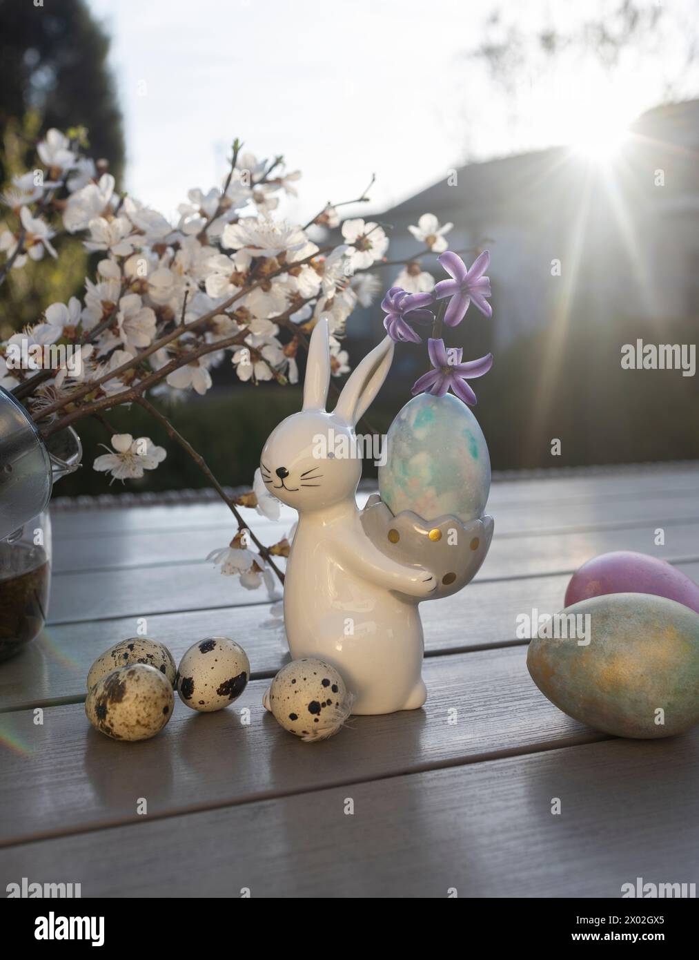 souvenir porcelain Easter bunny stands on the table in the rays of the setting back sun and holds an egg. spring white blossoming tree branches. Home Stock Photo