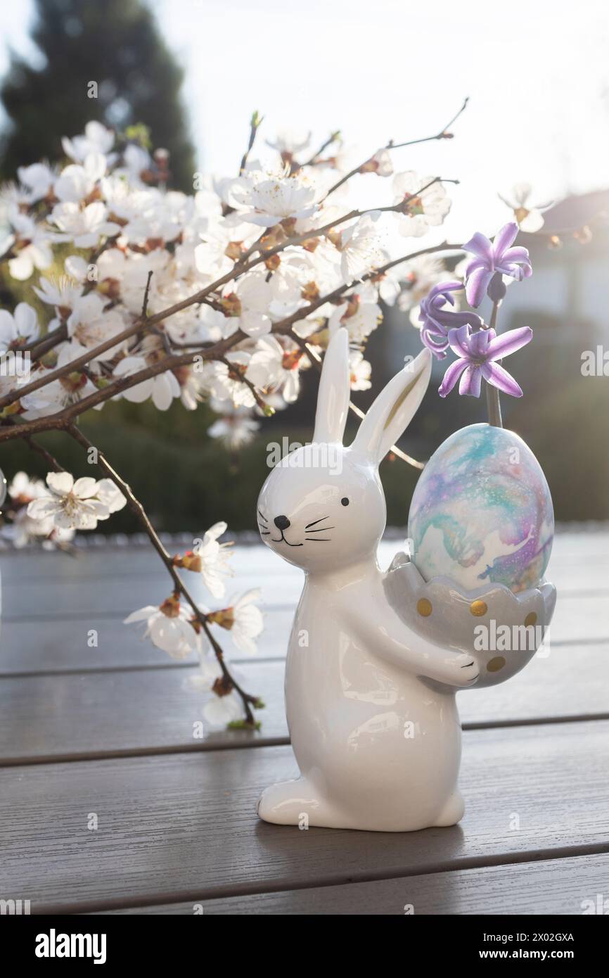 souvenir porcelain Easter bunny holds an egg against the backdrop of spring white tree branches blooming. Home holiday decor Stock Photo