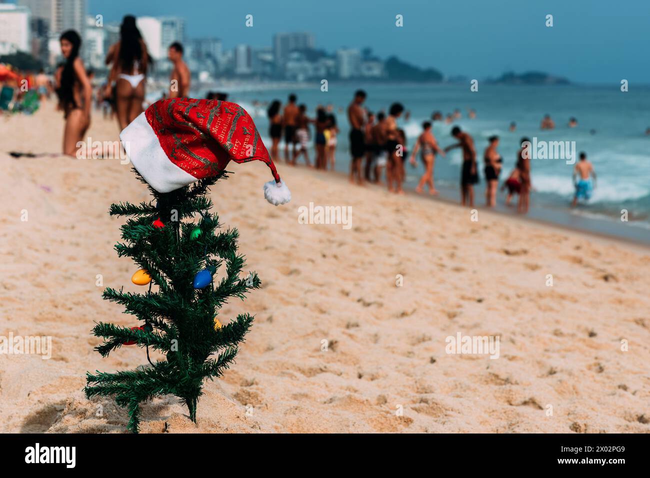 A makeshift small traditional Christmas tree and Santa Claus hat planted on the sand at Leblon Beach with a defocused crowd in the background Stock Photo