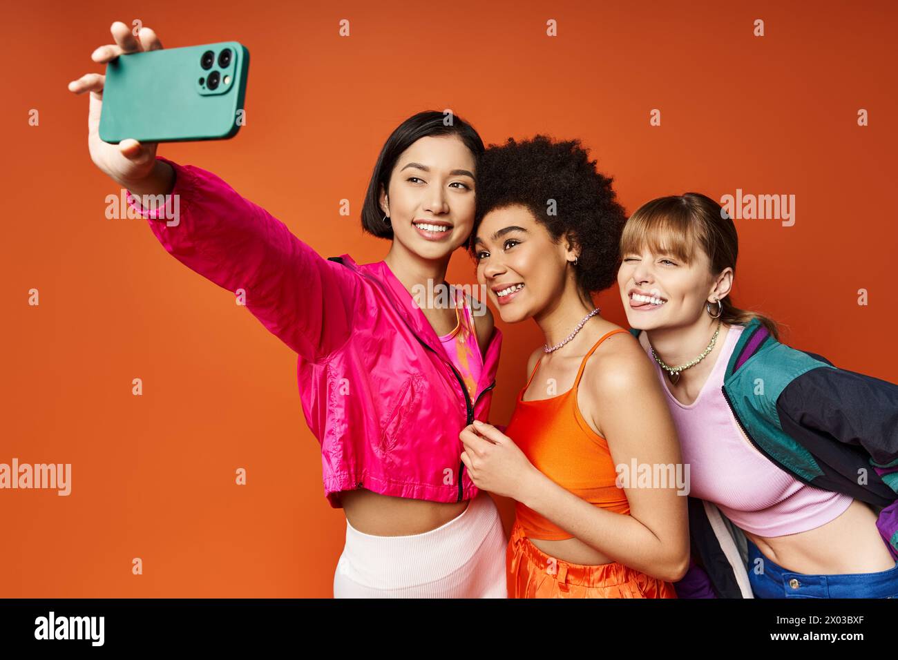 Three women, representing different cultures, enjoying a playful moment as they take a selfie with a cell phone against an orange background. Stock Photo